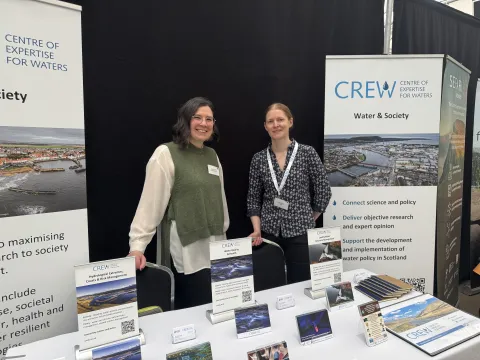 Image of Amy Cooper and Nikki Dodd from CREW behind the CREW stand at the ENRA conference. Postcards and leaflets are across a table in front of them and two pull ups stand behind them.