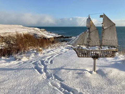 Snowy coastal landscape featuring a metal sailboat sculpture mounted on a pole in the foreground. The scene includes snow-covered ground with visible footprints, dry grass patches, rocky shoreline, and calm ocean under a partly cloudy blue sky.