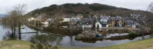 A village flooded by high water, with streets and buildings partially submerged, leafless trees standing in floodwater, and wooded hills in the background under a grey sky. Photo Credit: Ian McCourt - Callander Flood Group
