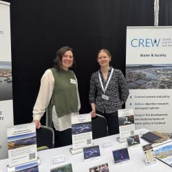 Image of Amy Cooper and Nikki Dodd from CREW behind the CREW stand at the ENRA conference. Postcards and leaflets are across a table in front of them and two pull ups stand behind them.