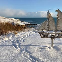 Snowy coastal landscape featuring a metal sailboat sculpture mounted on a pole in the foreground. The scene includes snow-covered ground with visible footprints, dry grass patches, rocky shoreline, and calm ocean under a partly cloudy blue sky.