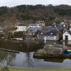 A village flooded by high water, with streets and buildings partially submerged, leafless trees standing in floodwater, and wooded hills in the background under a grey sky. Photo Credit: Ian McCourt - Callander Flood Group